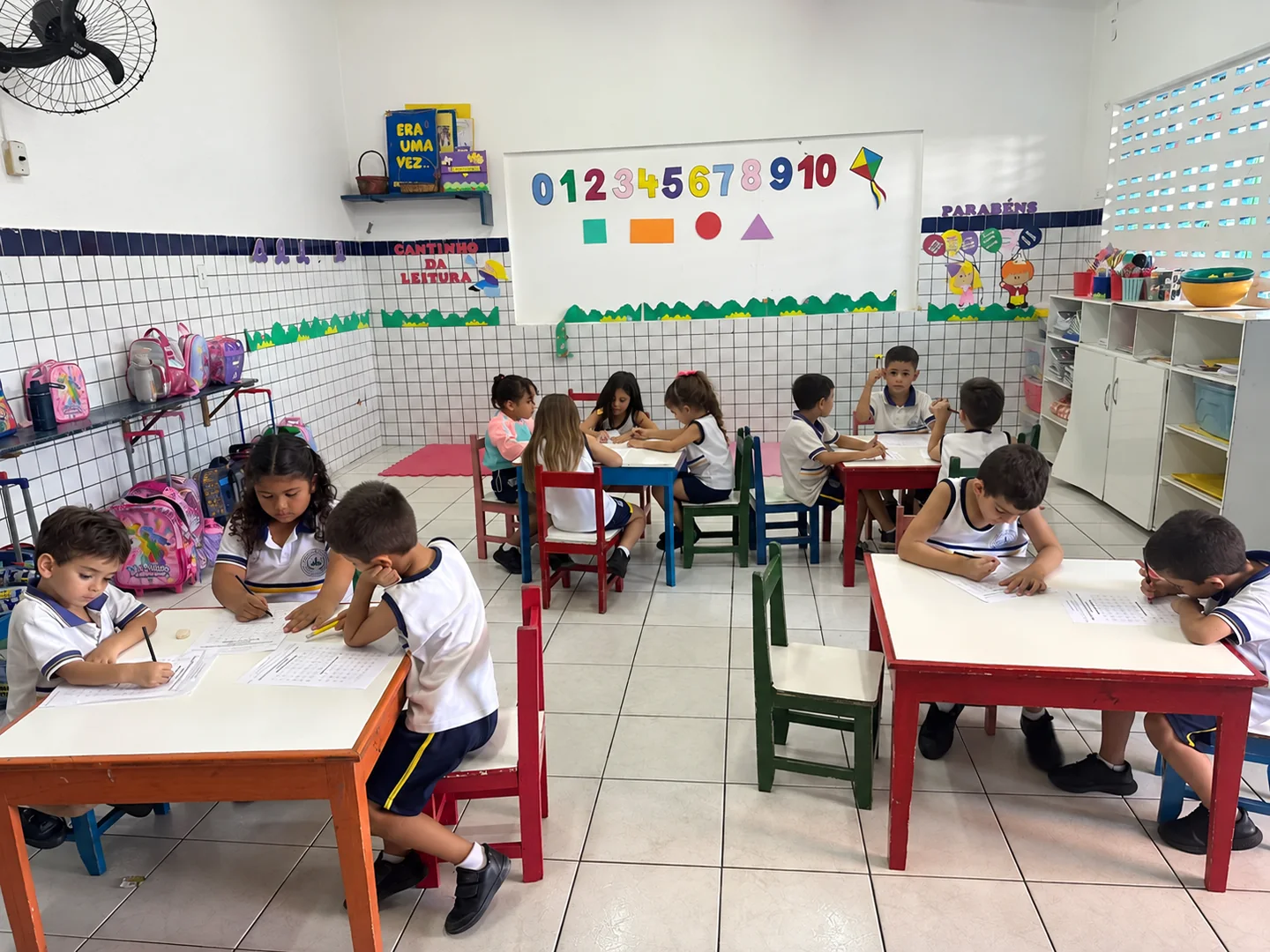 Sala de aula da Educação Infantil com crianças realizando atividades em mesas coloridas e materiais pedagógicos